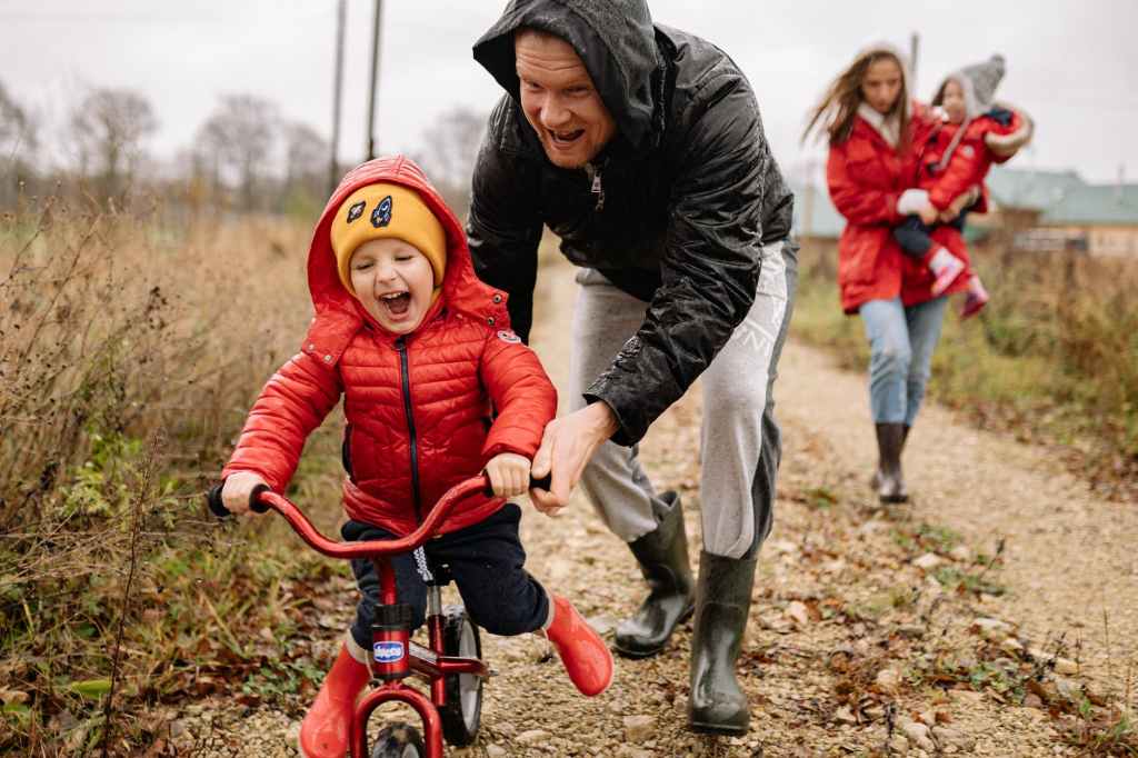 The image shows a father helping his toddler to ride a bike. 5 Heart-warming Reasons Why Performing Acts of Kindness is Good For You.